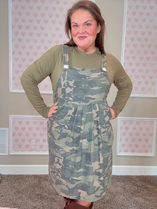Woman wearing a green long-sleeve top and camouflage overall dress in a room with pink heart-patterned wallpaper.
