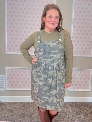 Person wearing a camouflage overall dress in a room with pink floral wallpaper