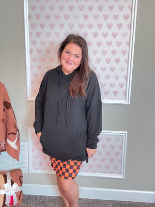 Person wearing a black hoodie and orange checkered skirt standing in front of a heart-patterned wall.