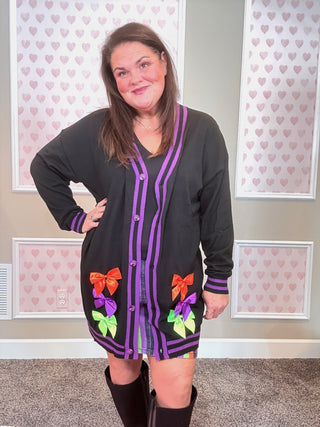Woman wearing a black cardigan with colorful bow designs in a room with heart-patterned wallpaper.