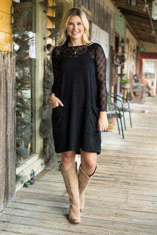 Woman in a black dress with sheer sleeves and beige boots standing on a wooden deck.