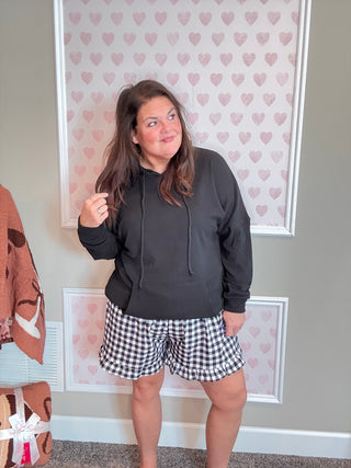 Woman wearing a black hoodie and checkered shorts standing in a room with heart-patterned wallpaper.