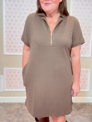 Woman wearing a brown dress with a zipper in a room with pink floral wallpaper.