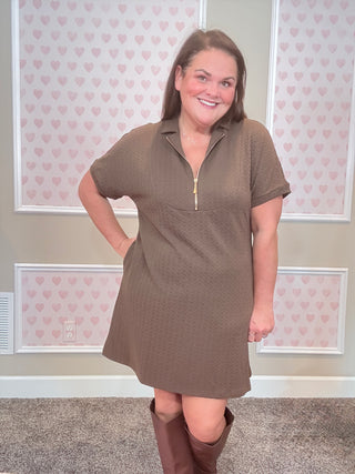 Woman wearing a brown dress standing in a room with heart-patterned wallpaper.