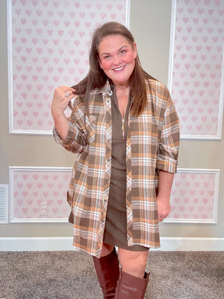 Woman wearing a plaid shirt over a dress with brown boots in a room with heart-patterned wallpaper.