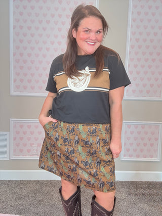 Woman wearing a graphic t-shirt and western patterned skirt with cowboy boots indoors.
