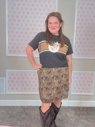 Woman wearing a graphic t-shirt and western patterned skirt with cowboy boots in a room with heart-patterned wallpaper.