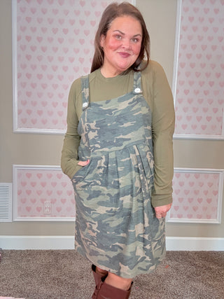 Person wearing a camouflage overall dress in a room with pink heart wallpaper.