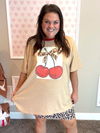 Person wearing a beige t-shirt with a graphic design of cherries and a bow, standing in a room with heart-patterned wallpaper.