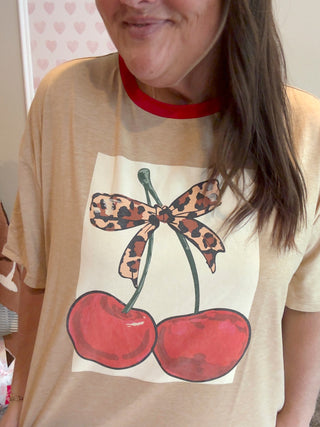 Person wearing a beige t-shirt with a graphic of cherries and a leopard print bow.