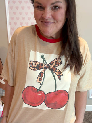 Person wearing a beige t-shirt with a graphic of cherries and a leopard print bow.