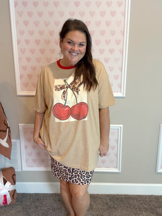 Woman wearing a beige t-shirt with a graphic cherry design and leopard bow and leopard print skirt in a room with heart-patterned wallpaper.