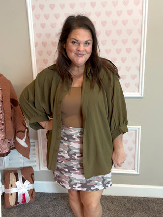Woman wearing a green top, brown top, and camouflage skirt in a room with heart-patterned wallpaper.