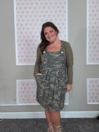 Woman wearing a camouflage dress over a green top in front of a wall with heart patterns.
