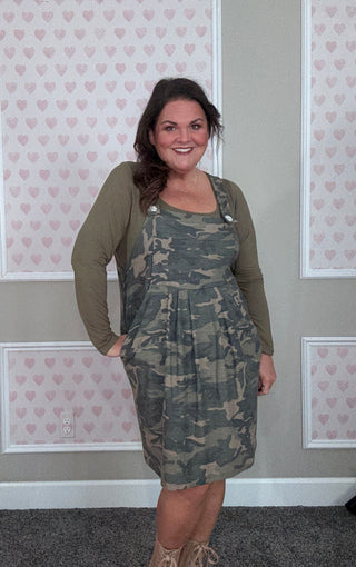 Woman wearing a green camouflage dress with a green top in front of a wall with heart patterns.