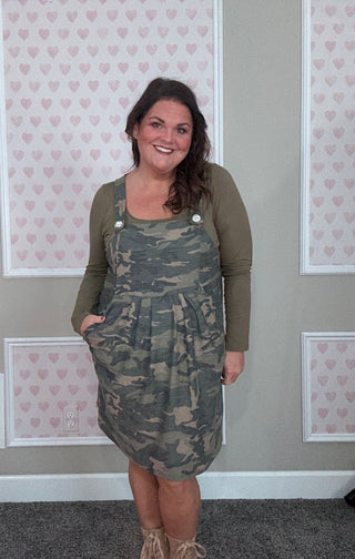 Woman wearing a camouflage dress with a green top in front of a wall with heart patterns.