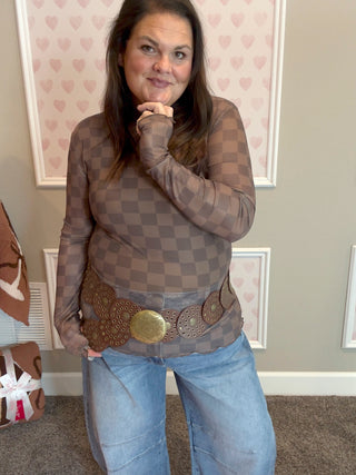 Woman wearing a sheer checkered top with a wide belt in a room with heart-patterned wallpaper.