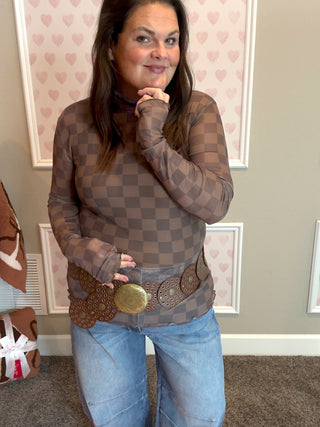 Woman wearing a sheer checkered top and jeans indoors with decorative wall paper.