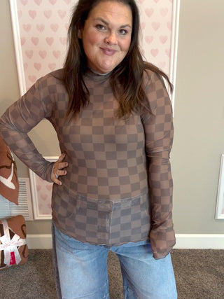 Person wearing a brown checkered top and jeans indoors with a heart-patterned wallpaper.