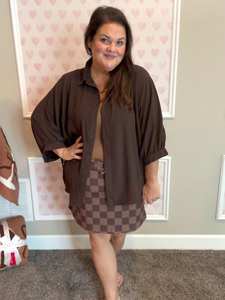 Woman wearing a brown shirt and checkered skirt standing in a room with heart-patterned wallpaper.