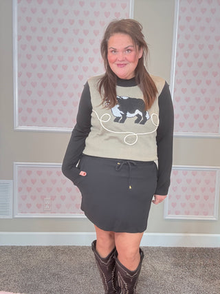 Woman wearing a beige sweater with a graphic design and a black skirt in a room with heart-patterned wallpaper.