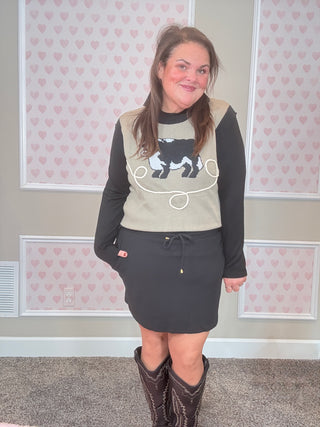 Woman wearing a graphic sweater and skirt with cowboy boots in a room with heart-patterned wallpaper.