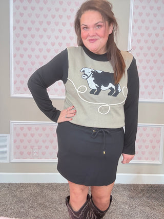 Woman wearing a sweater with a cow design in a room with heart-patterned wallpaper.