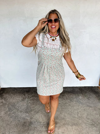 Woman wearing a floral dress and sunglasses against a white wall.