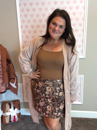 Woman wearing a beige cardigan, brown top, and floral skirt in a room with heart-patterned wallpaper.