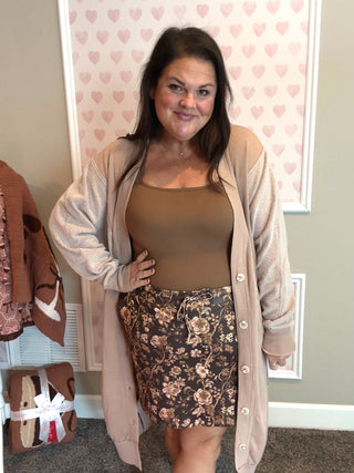 Woman wearing a beige cardigan, brown top, and floral skirt in a room with heart-patterned wallpaper.