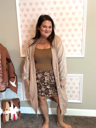 Woman wearing a beige cardigan, brown top, and floral skirt in a room with heart-patterned wallpaper.