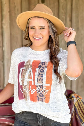 Woman wearing a graphic t-shirt with 'Vibes' and a hat, standing against a wooden wall.