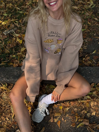 Person wearing a beige sweatshirt with a graphic design sitting on a leaf-covered ground.