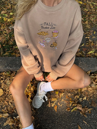 Person wearing a beige sweatshirt with 'Fall Bucket List' design sitting on leaves.