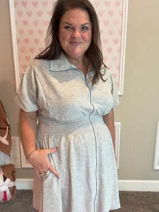 Woman wearing a light gray dress standing in a room with heart-patterned wallpaper.