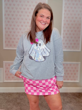 Woman wearing a gray sweatshirt with a ghost design and a pink checkered skirt in a room with heart-patterned wallpaper.