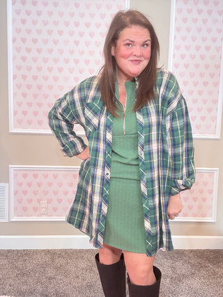 Person wearing a green and blue plaid flannel over green dress with a patterned wall in the background