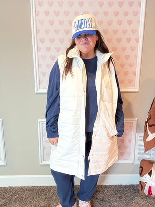 Woman wearing a long cream puffer vest over navy jogger outfit with a 'GAMEDAY' cap, standing in a room with heart-patterned wallpaper.
