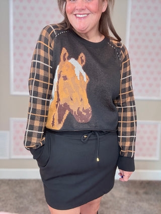 Person wearing a sweater with a horse design and plaid sleeves in a room with pink walls.
