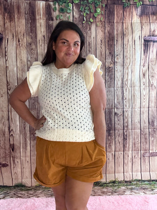 Woman wearing a white top with ruffled sleeves and mustard shorts against a wooden fence background.