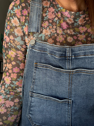Person wearing a floral shirt and denim overalls with a focus on the overalls.