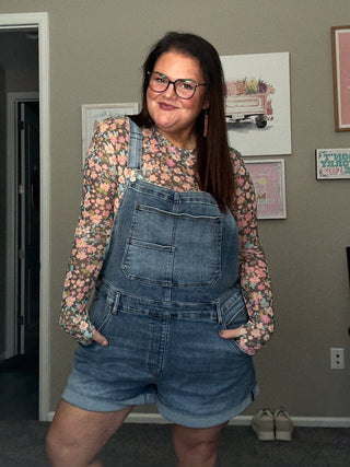 Woman wearing denim overalls and a floral shirt in a room with framed pictures on the wall.