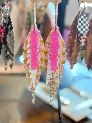 Layered faux leather earrings featuring hot pink glitter on top, yellow bow print, and a lined notebook paper pattern with silver chain accents. Fun, colorful, and school-themed statement earrings displayed on a “randans” earring card.