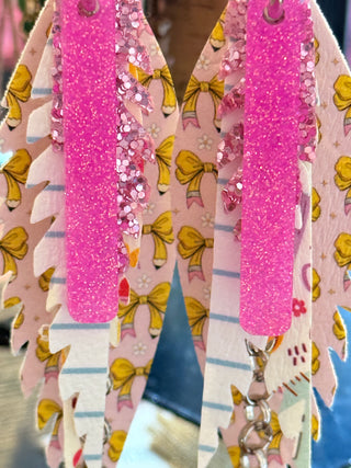 Layered faux leather earrings featuring hot pink glitter on top, yellow bow print, and a lined notebook paper pattern with silver chain accents. Fun, colorful, and school-themed statement earrings displayed on a “randans” earring card.