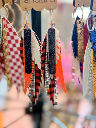 Handmade tiered earrings with layers of orange, black glitter, white leather, and black-and-orange buffalo plaid, hanging from silver hoops on a "randans" card.