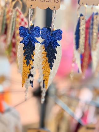 Tiered leather earrings in blue, gold, and white with glittery blue bows on top. Features golden paisley, navy floral, and sparkly white layers with silver chain dangles. Displayed on a “randans” card with silver hoops.
