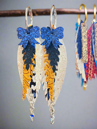 Tiered leather earrings in blue, gold, and white with glittery blue bows on top. Features golden paisley, navy floral, and sparkly white layers with silver chain dangles. Displayed on a “randans” card with silver hoops.