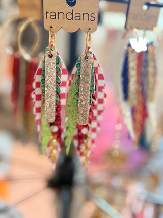 Layered holiday earrings with red-and-white checkerboard, green glitter, and red sparkle feathers. Topped with a gold glitter bar and finished with gold chain fringe, hanging from gold hoops. Displayed on a “randans” earring card.