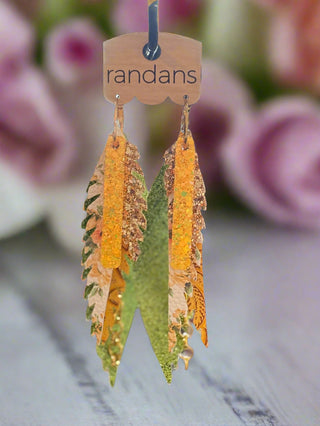 A pair of tiered feather earrings featuring green suede, gold glitter, and floral faux leather layers with gold chain accents, hanging from stainless steel hoops on a branded “randans” earring card. Displayed on a jewelry rack.