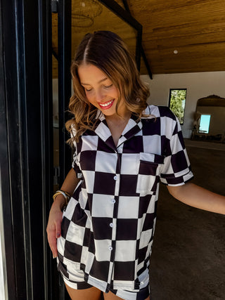 Model lounging in a black and white checker luxe pajama set with button-up top and shorts.